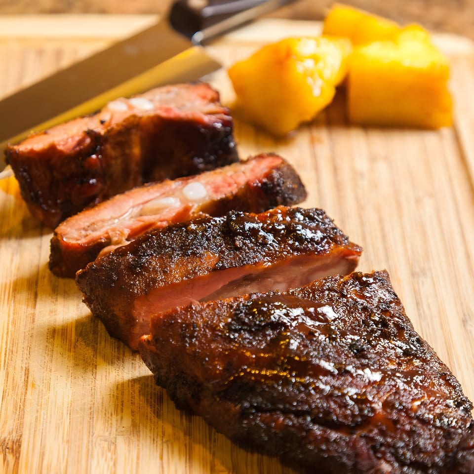 Close-up of Bill's Grill Smokehouse BBQ ribs, sliced on a wooden cutting board with pineapple.